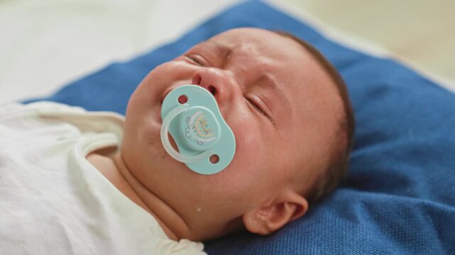 Hispanic baby boy crying, held by adult hands, pacifier visible, tears on cheeks, crib rails and bedding visible in a building; distress comfort bonding.