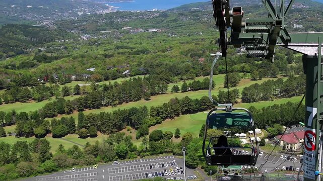 Scenic horizontal video from Mount Omuro in Izu, Japan, with a chairlift descending over a grassy hillside toward forest, town, coastline and blue sea. Peaceful summer travel and landscape concept.