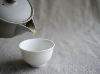 Obraz premium Pouring fresh hot green tea from a gray ceramic teapot into a white porcelain cup on fabric background