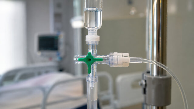Clear plastic infusion set connector green stopcock valve hospital equipment close up with tube, fluid chamber, pole, monitor, sterile clinical detail, blurred room background