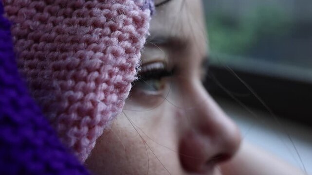 Intimate close-up shot of a child wrapped in a textured pink and purple knitted scarf, quietly gazing out a window with soft daylight casting gentle shadows on their face.