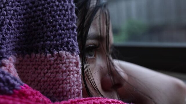 A thoughtful young girl wrapped in a knitted purple and pink blanket gazes pensively out a window on a moody, rainy day, captured in a soft close-up shot with natural light.