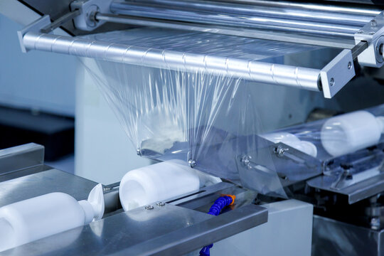 automatic shrink wrapping machine applying clear plastic film to white lotion cream pump bottles on conveyor belt in cosmetic factory. Industrial packaging, processing technology, and manufacturing.