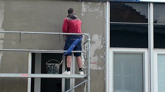 Construction worker applying plaster to building exterior from scaffolding. A man on scaffolding uses a trowel to apply plaster to a building facade near windows.