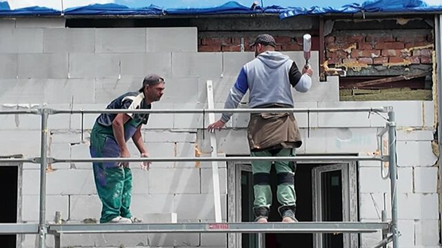 Construction workers building a wall with concrete blocks on scaffolding. Skilled laborers meticulously construct a building facade using lightweight concrete blocks and scaffolding for elevated