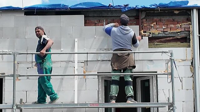 Construction workers building a wall with concrete blocks on scaffolding. Workers meticulously place concrete blocks, constructing a new wall on a building site, showcasing the process of modern