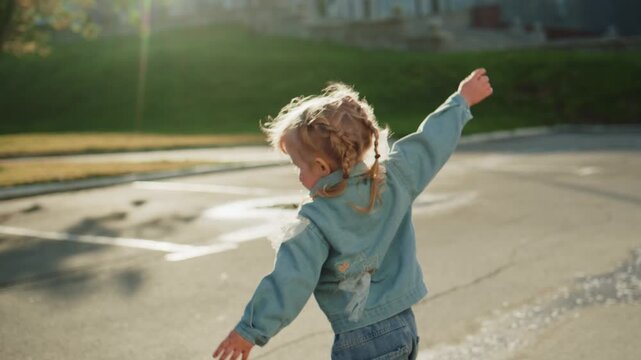 Little girl plays in puddles on sunny day. She runs joyfully with arms outstretched. Sunlight highlights her curly hair and denim jacket