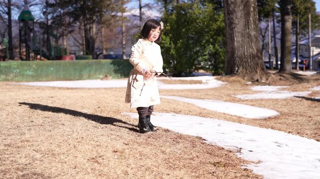 Soft sunlight warms winter's last snow patches. Child in a long coat walks through melting ground. She bends low, curious, to touch the earth's last chill