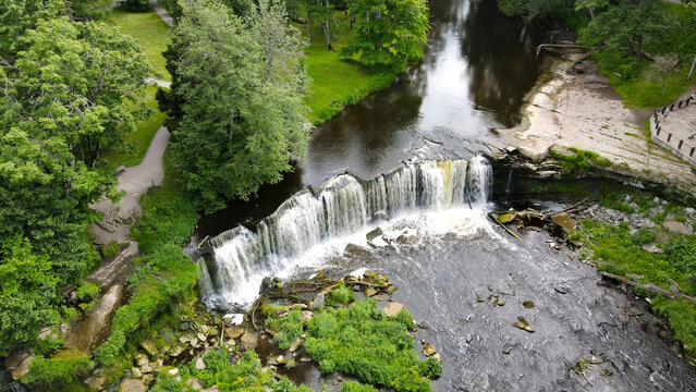 Keila waterfall displaying water cascading over a wide rock formation, surrounded by lush green trees and vegetation along the riverbank in a peaceful natural environment