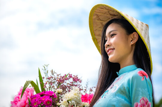 Pretty young Vietnamese woman in traditional Ao dai and hat holding flowers in nature 