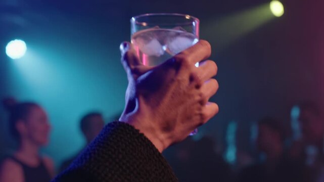 Close up of a hand raising a glass of refreshing cocktail with ice at a crowded nightclub party with colorful strobe lights