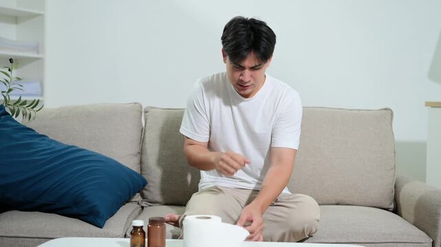 A sick young Asian man experiencing flu symptoms, coughing and using a tissue while sitting on a couch at home.