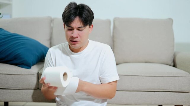 A sick young Asian man experiencing flu symptoms, coughing and using a tissue while sitting on a couch at home.