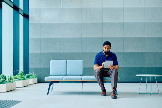 Black man sitting on a waiting room bench, using a tablet for connectivity, business, and entertainment in a modern office foyer