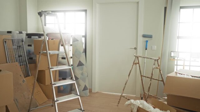 Two women hold cardboard boxes at waist, smiling while lifting near ladder and window during moving in building; friendship joy.