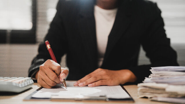 Professional hand writes document at desk. Focused person signs contract with red pen. Stacked papers nearby suggest busy workload. Office setting conveys serious business tone