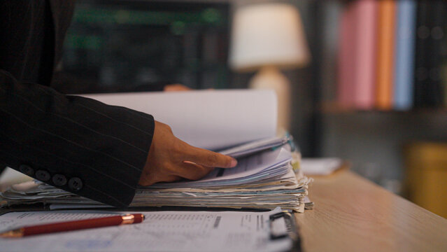 Hand flips through thick paperwork on a wooden desk. Soft light glows from a lamp in the background. Piled documents suggest heavy workload or deadline pressure. Person wears dark jacket, focused