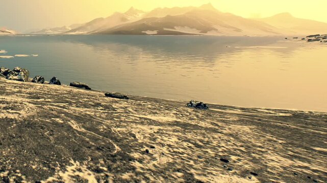 A rocky shoreline is visible with stones on the ground. In the background, mountains rise against a sunset sky. Water reflects colors from the sky creating a beautiful scene.
