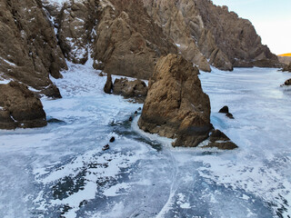 Aerial view of expedition vehicles (appearing as dark objects) moving across the textured, frozen surface of the gorge past a massive boulder and sheer rock walls © Collab Media