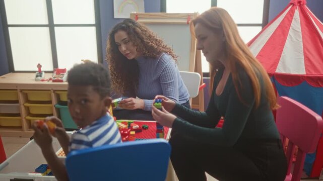 Women help boy stack colorful blocks at a small table with tent and storage drawers in a classroom inside building; playful learning joy.