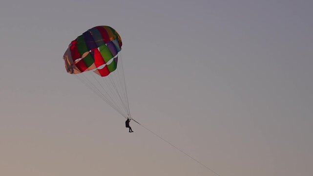 Thrilling parasailing adventure with a single person harnessed to a vibrant, multicolored parachute, gliding gracefully across a clear, minimalist sky for an exciting recreational sport