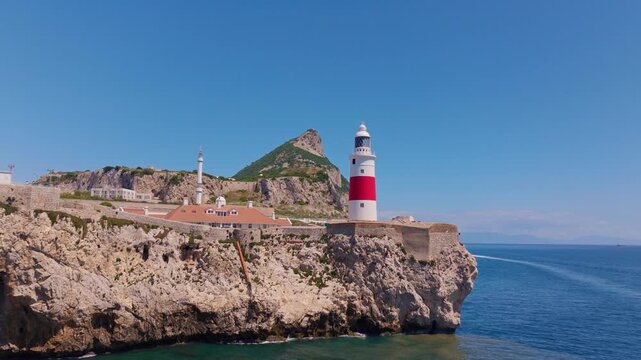 Drone footage approaching and ascending past Europa Point lighthouse &mdash; red and white tower, Ibrahim-al-Ibrahim Mosque, limestone cliffs, and Mediterranean Strait beyond.