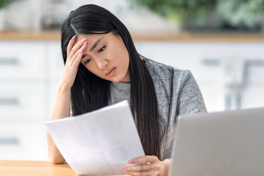Stressed Asian woman reviewing financial paperwork laptop