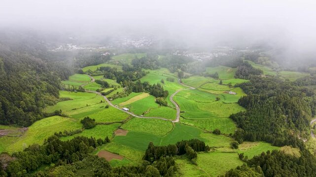 Cinematic drone aerial view of the lush green countryside on Sao Miguel Island Azores Portugal featuring patchwork fields winding rural roads dense forests and low clouds drifting across the landscape