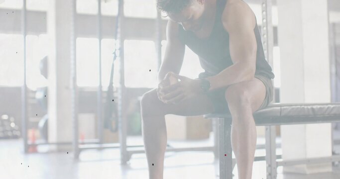 Settling man in tank and shorts resting between sets on bench at gym, leaning, clasping hands