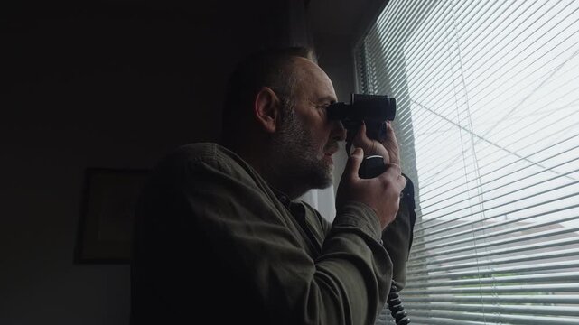 : An undercover observer scans through binoculars behind window blinds while transmitting updates by radio. Strong espionage, surveillance, intelligence, and thriller storytelling concept.
