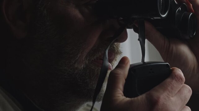 Close-up of a bearded operative quietly reporting observations over a handheld radio while watching through a window. Perfect for espionage, surveillance, detective, and crime thriller narratives.