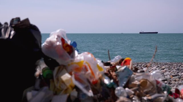 Plastic waste and other rubbish piled up on a pebble beach, contaminating the shoreline while a cargo ship sails on the distant sea, symbolizing a serious environmental problem and pollution