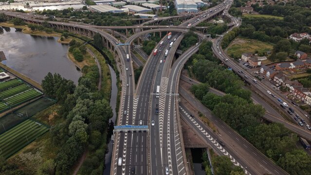 Aerial drone view looking along the M6 motorway through the famous Spaghetti Junction in Birmingham, England. Multiple elevated flyovers, heavy traffic, canals, surrounding industrial estates and urba