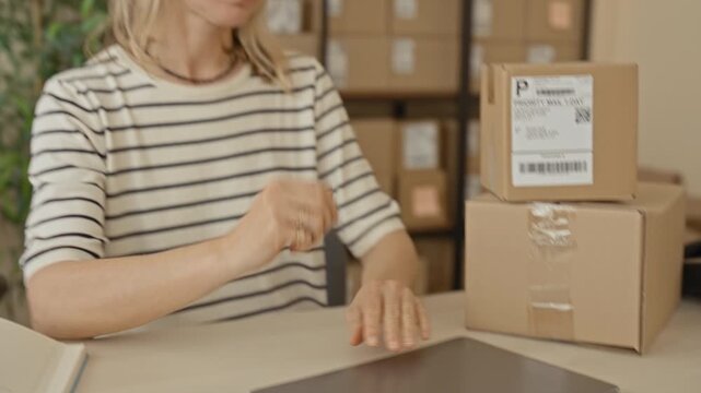 Woman holding parcel and rubbing eyes while touching glasses in a building office with stacked boxes and a laptop visible; small business stress tired.