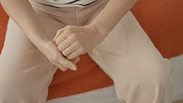 Woman seated on bed with hands clasped and rubbing fingers, fidgeting and massaging knuckles in bedroom; anxiety introspection.