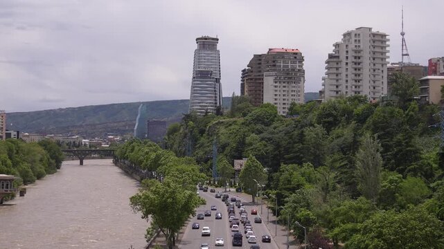 Aerial view of cars and vans driving on a busy highway along the embankment of the muddy mtkvari river in tbilisi, the capital of georgia, surrounded by lush green trees and city buildings