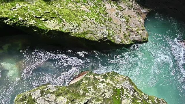 Top view of a stream of clear turquoise water flowing through a narrow rocky gorge with bright green, moss-covered shores.
