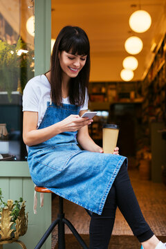 Smiling woman sitting on stool using cell phone at entrance door of a store