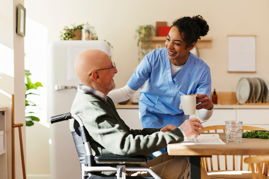 Smiling caretaker giving mug to senior man on wheelchair at home