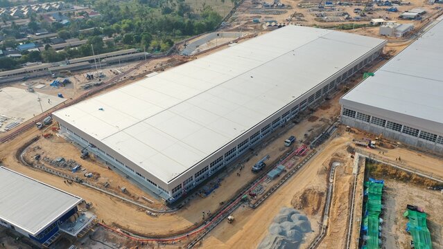 Construction of a large industrial warehouse under clear sky, showing modern architecture and industrial development, with surrounding infrastructure and ongoing building work.