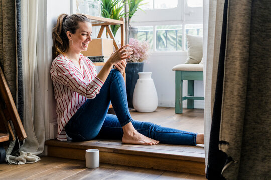 Smiling woman sitting on the floor at home with cell phone