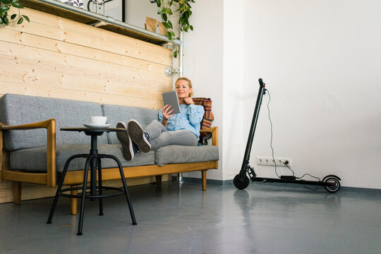 Young woman sitting on couch in a coffe shop, using digital tablet, e-scooter charging in background