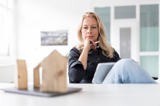 Thoughtful businesswoman with digital tablet looking at model on desk while sitting in home office
