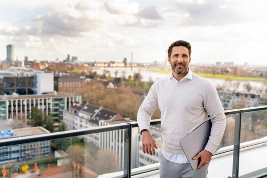 Smiling male entrepreneur with laptop leaning on railing at balcony in office