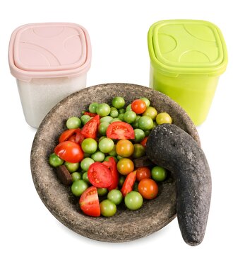 Traditional Indonesian stone mortar and pestle with fresh red and green tomatoes, red chili peppers, and plastic kitchen containers on white background