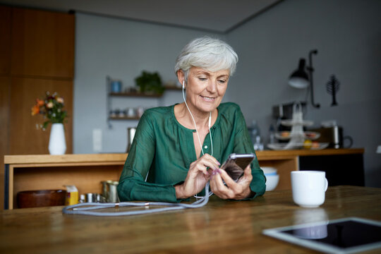 Senior woman text messaging and listening music on smart phone while sitting at home