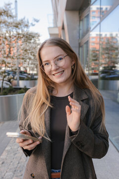 Smiling businesswoman waving and holding smartphone outdoors