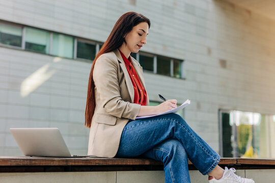 Businesswoman in jacket sitting outdoors writing on notepad