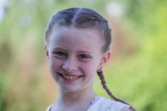Portrait of smiling young girl with braided hair outdoors. Closeup portrait of happy caucasian kid with blue eyes and braids smiling in sunny summer park