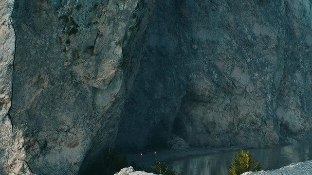 Two climbers navigate the rocky area next to a river. The sun shines down, creating clear visibility for their activity. The natural surroundings are rugged and wild.